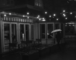 Õhtune stseen Pärnu kesklinnas: mustvalge jäädvustus daamist, kes seisab vihmavarju all terrassikohviku valgusketis. Autor: Priit Suve. Mustvalge Priit Suve tänavafoto Pärnu kesklinnast: daam vihmavarjuga seisab õhtul kohviku sissepääsu ees, mille ees on terrass ja tuled.