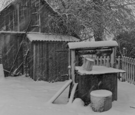Talvine kaev Lääne-Virumaal. Lumi ja tuisk katavad vana kaevu ning selle ümbruse, rõhutades maastiku vaikust ja ajatut rütmi. Mustvalge talvine kaev Lääne-Virumaal lumesajus, vana puithoone ja aiaga taustal